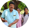 A man in a light blue polo shirt holding papers, with two young women standing behind him outdoors under a tree.